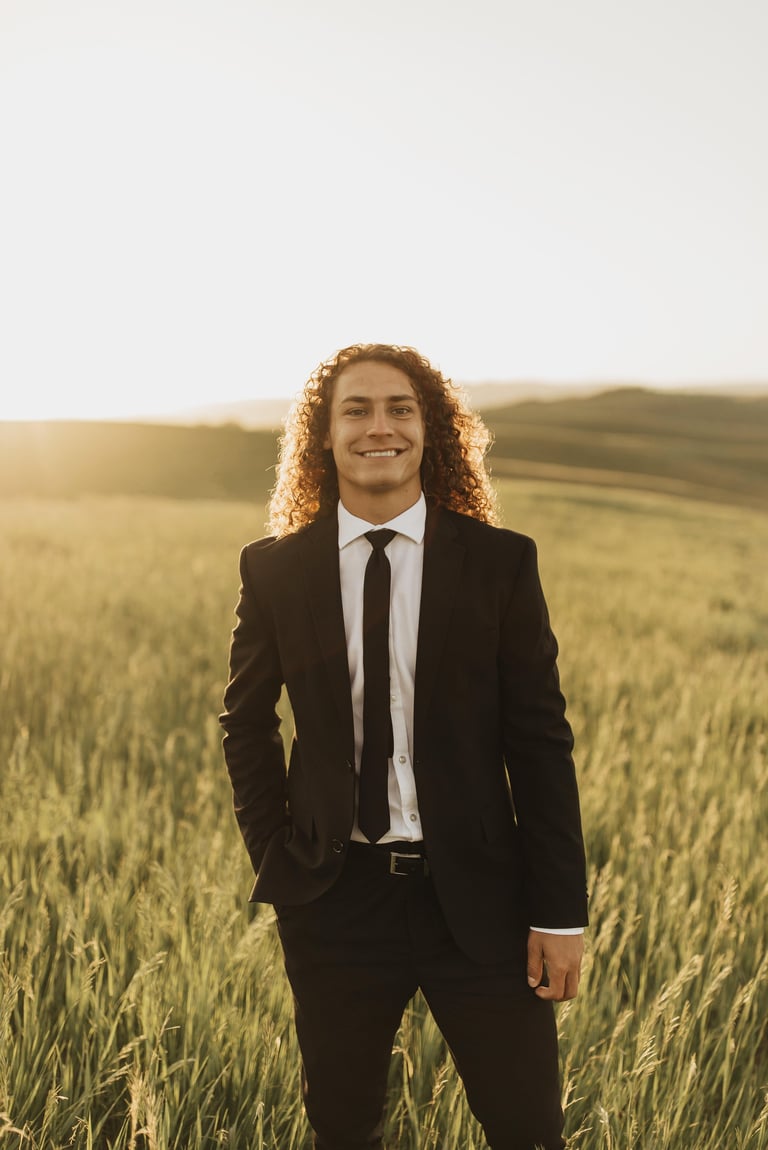 Man with curly hair in black business suit and tie standing in golden grain field at sunset